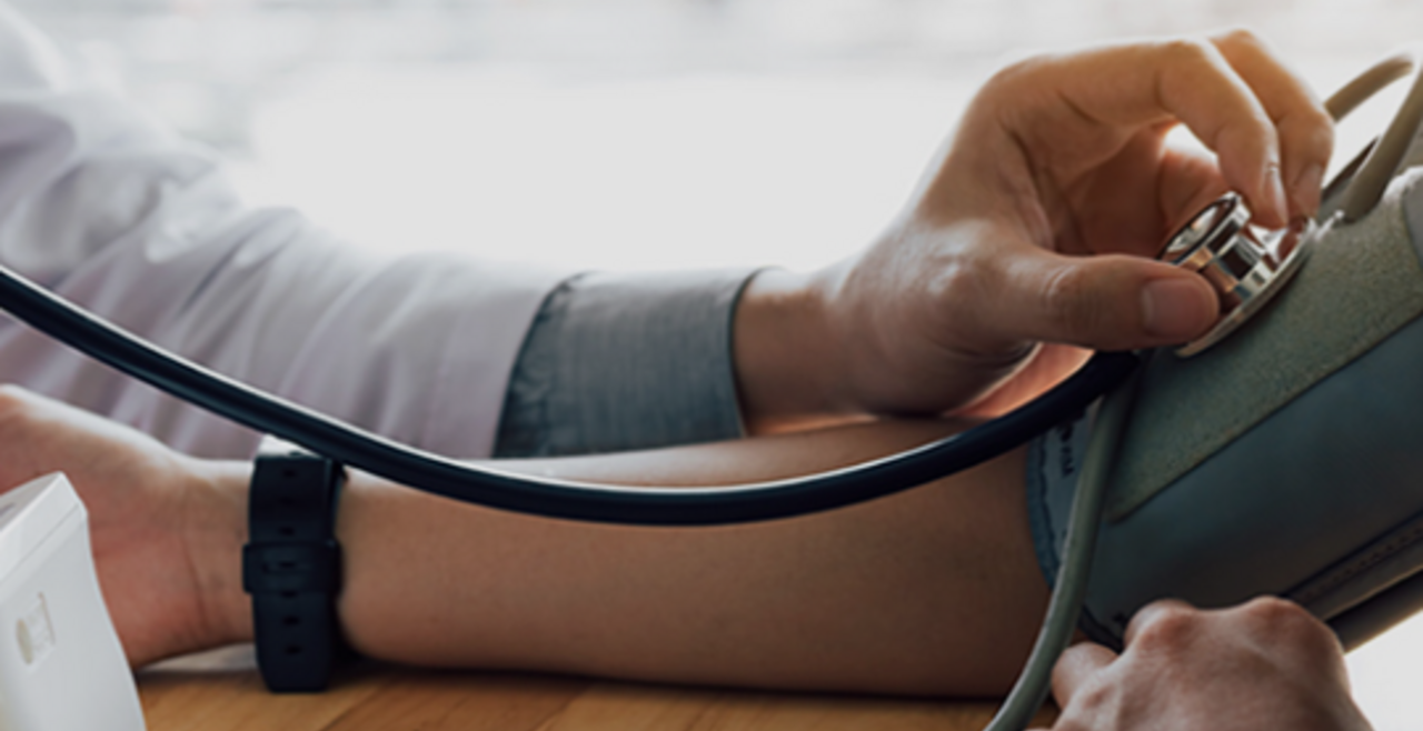 A close-up on a black individual’s arm with a pressure cuff, and a doctor’s hand placing a stethoscope on the cuff.